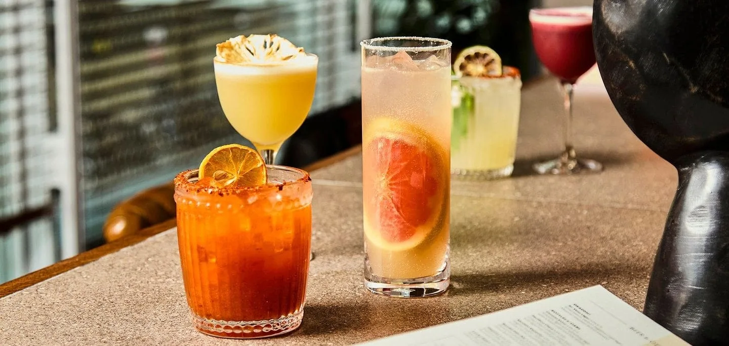 A selection of colourful, refreshing Mexican inspired and tequila based cocktails sitting on the bar. The grapefruit paloma, the marmalade mezcalita and the pineapple mezcal sour. The mezcal sour is a vegan cocktail due to the use of aquafaba from chickpeas instead of using egg whites.