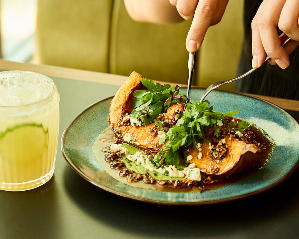 Someone cuts into a large Mexican inspired vegetarian dish - roasted organic squash, served with a cashew nut and fresh herb sauce and nutty salsa macha. it is garnished with coriander and feta, though the dish can be made vegan. A classic margarita cocktail sits by the side of the dish.