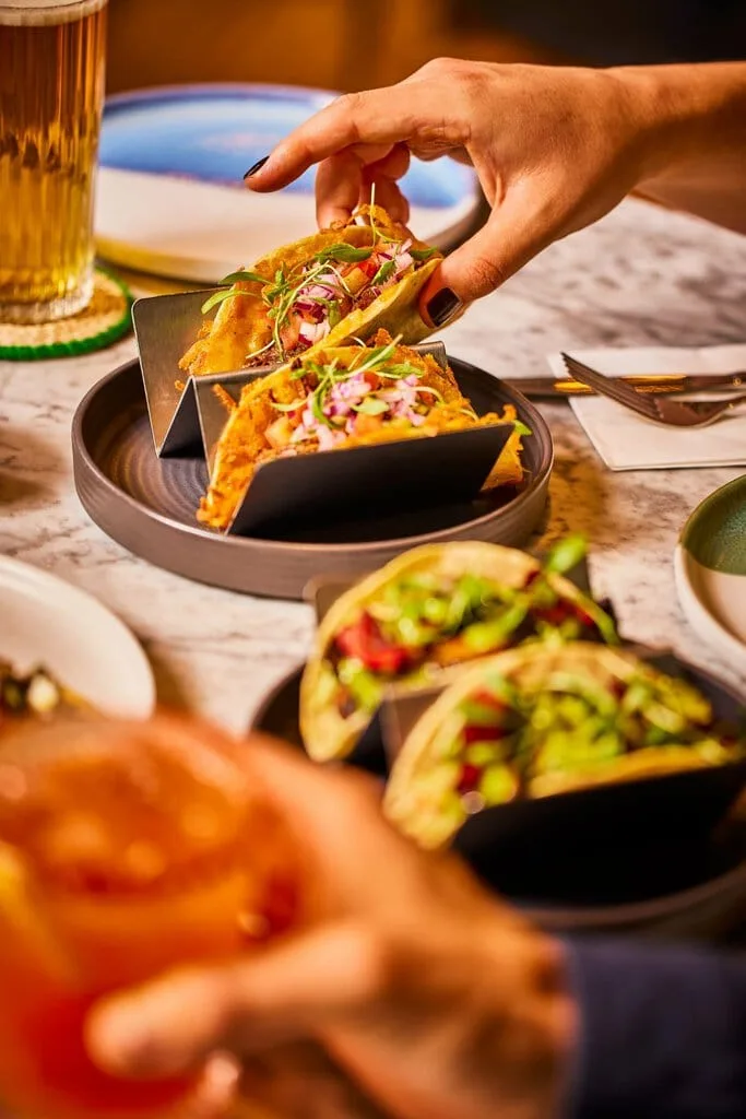 A woman picks up a beef gringa taco, one of our most popular Mexican dishes, in our Mexican restaurant in Paddington, London. The beef gringa tacos are made with a grilled cheese tortilla, pulled beef, fresh salsa and micro herbs. In the foreground, somebody holds a marmalade mezcal cocktail and our vegan ancho mushroom tacos sit on a dish on the table.