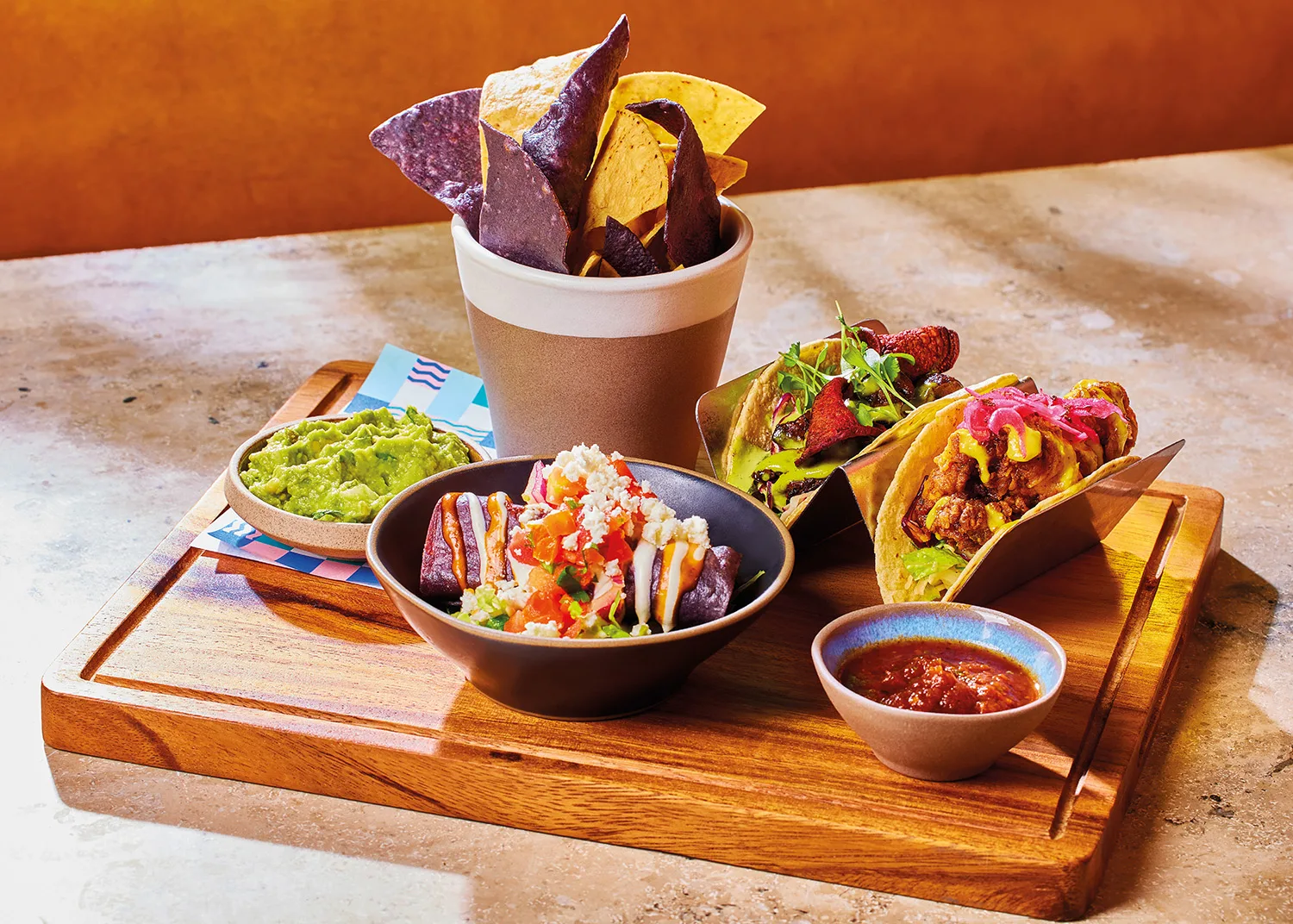 A selection of different Mexican dishes, including two tacos, a sweet potato and feta taquito, a pot of guacamole, a pot of smoky sonoran hummus and some tortilla chips, served on a board in our Mexican restaurant.