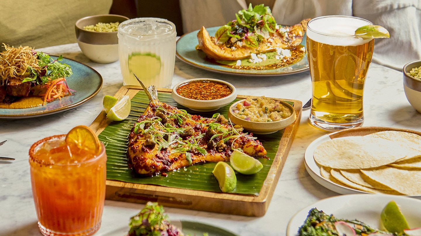 A table is filled with Mexican inspired plates of food in our Mexican restaurant. A whole grilled fillet of marinated seabass is serve on a banana leaf , with pots of charred pineapple salsa and nutty salsa macha, with 4 lime wedges. There is a plate of soft corn tortillas. There is a vegan food item, the organic roasted squash, served with cashew nut and herb mole sauce and salsa macha. There is a smoky caramelised pork belly dish, covered in tamarind sauce with crispy leeks served on top. Drinks on the table include a pint of carbon negative lager beer, freshly shaken marmalade mezcalita and a classic margarita cocktail.