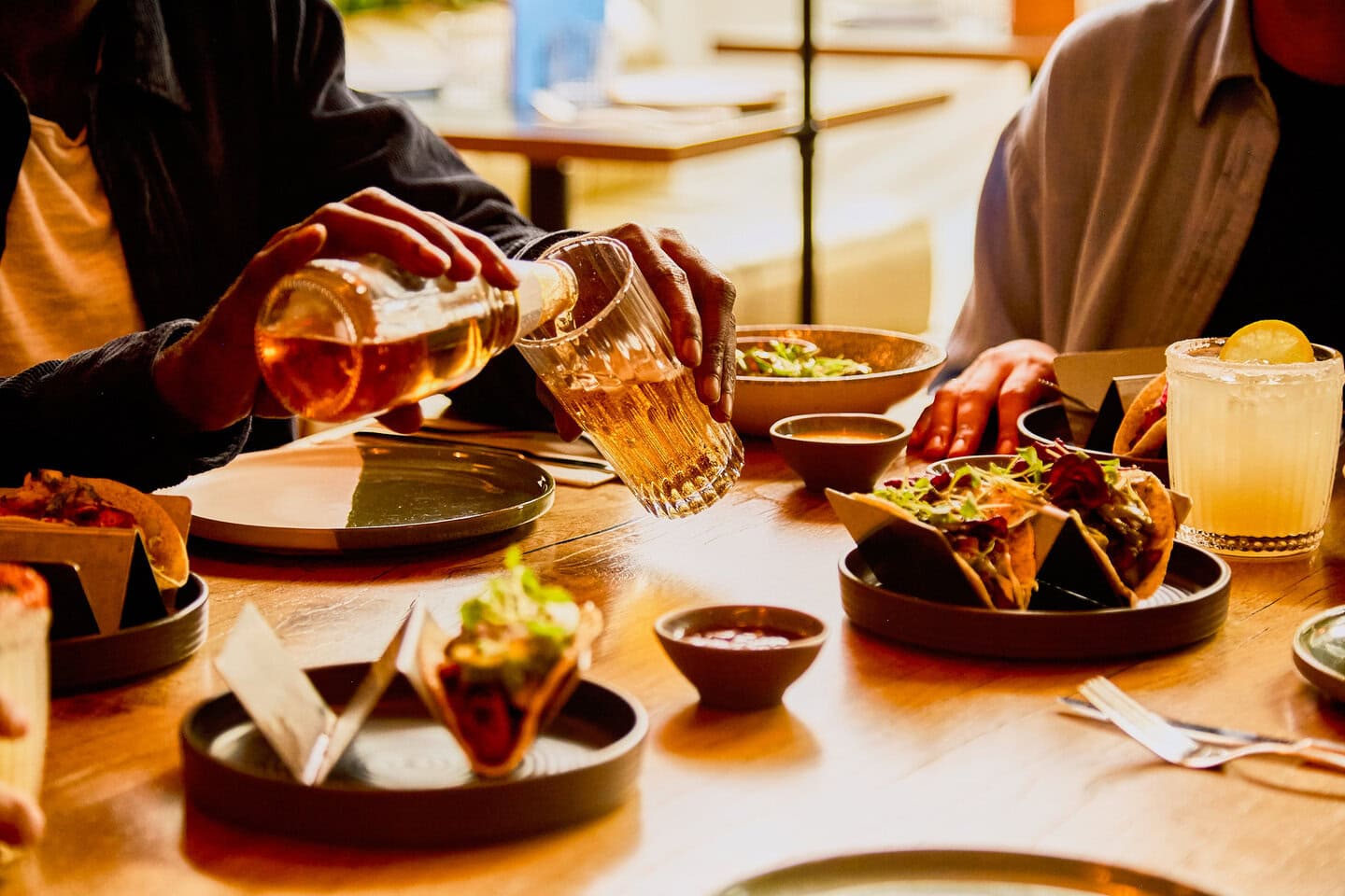 Two people sit at a table in our London restaurant that is filled with Mexican food, including vegan ancho mushroom tacos, free-range pork pibil tacos, vegetarian halloumi al pastor tacos, a Mexican lager beer and a classic margarita cocktail.