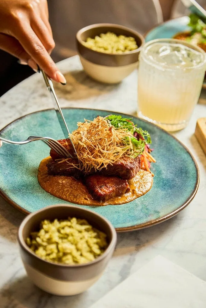 A large plate containing smoky caramelised pork belly that sits on top of tamarind sauce with a crisp leek garnish. Someone digs into the pork belly with their knife and fork.