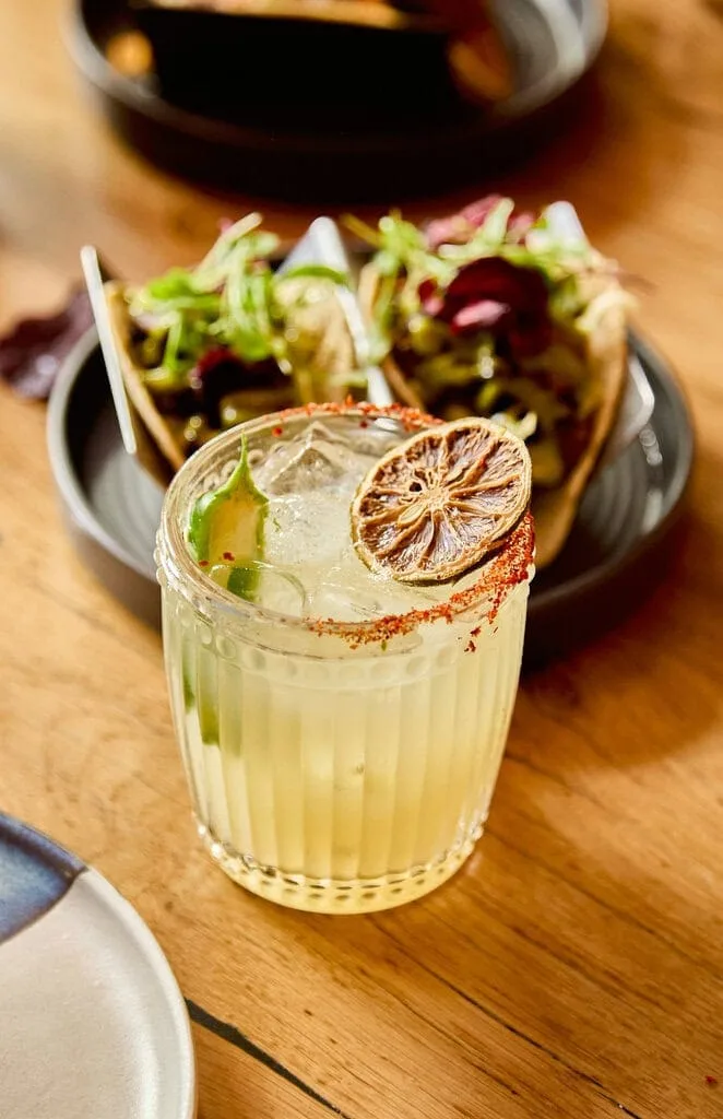 A refreshing picante margarita cocktail sits on a table in front of some vegetarian mushroom tacos. The margarita cocktail has a chilli salt rim and is served with half a green chilli and a dried lime slice