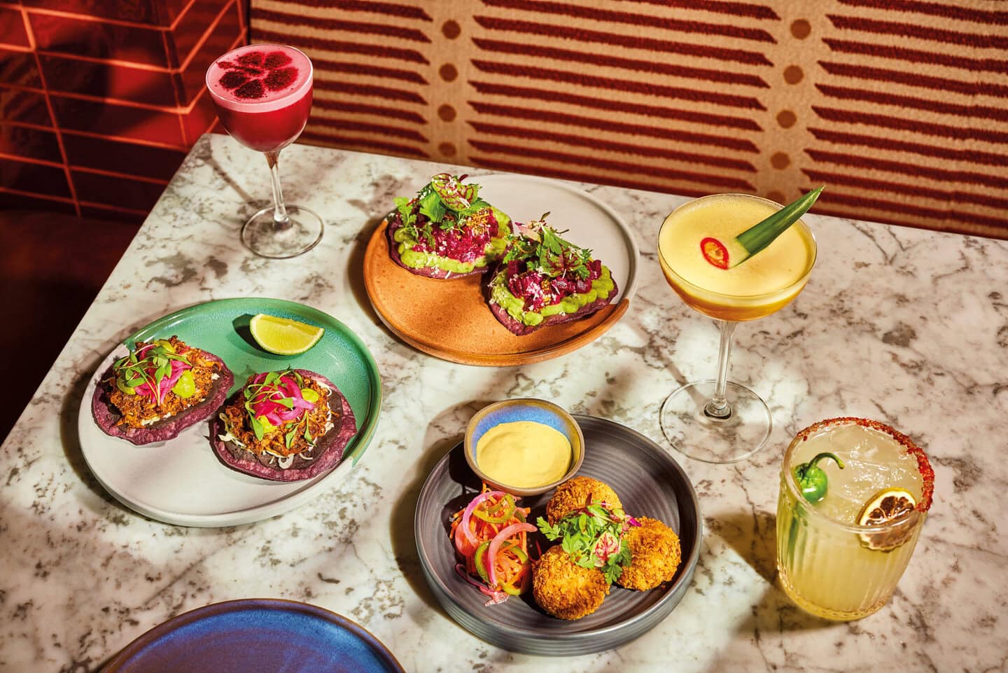 A restaurant table filled with colourful Mexican food and cocktails. Mexican food includes vegan beetroot tostadas and vegan mushroom carnitas, with regenerative beef croquetas. Mocktail selection includes alcohol free margarita, pineapple picante and and blackberry sour.